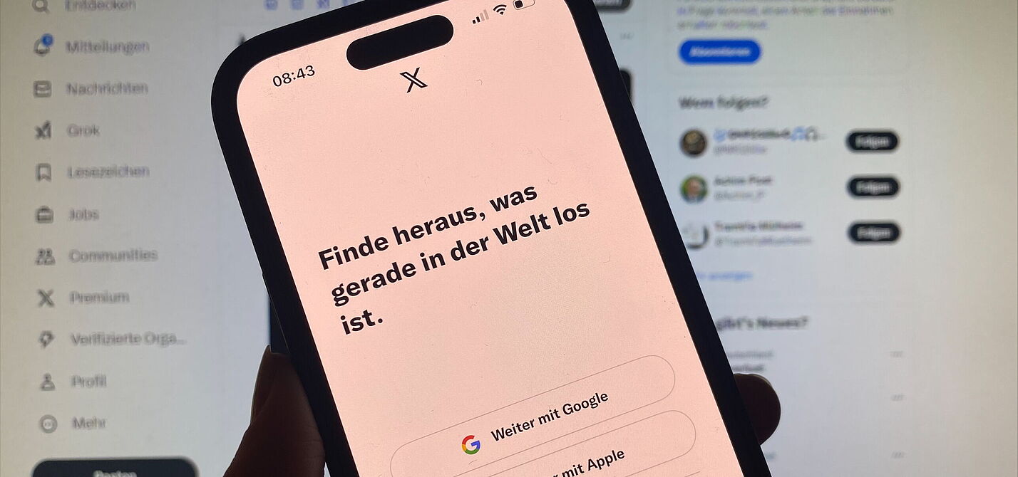 Ein Smartphone mit der geöffneten App "X" vor einem Computer-Bildschirm