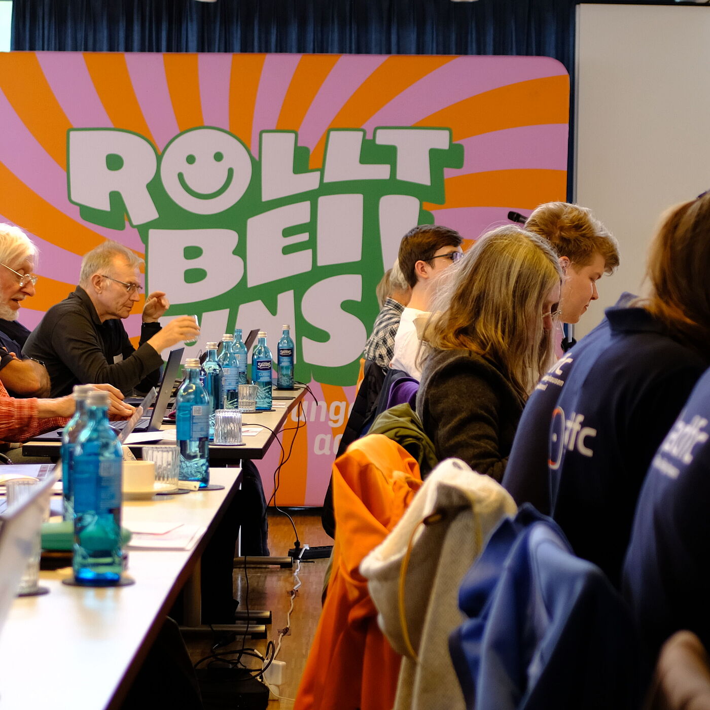 Mehrere Personen, Stuhlreihen mit Plakat im Hintergrund. 