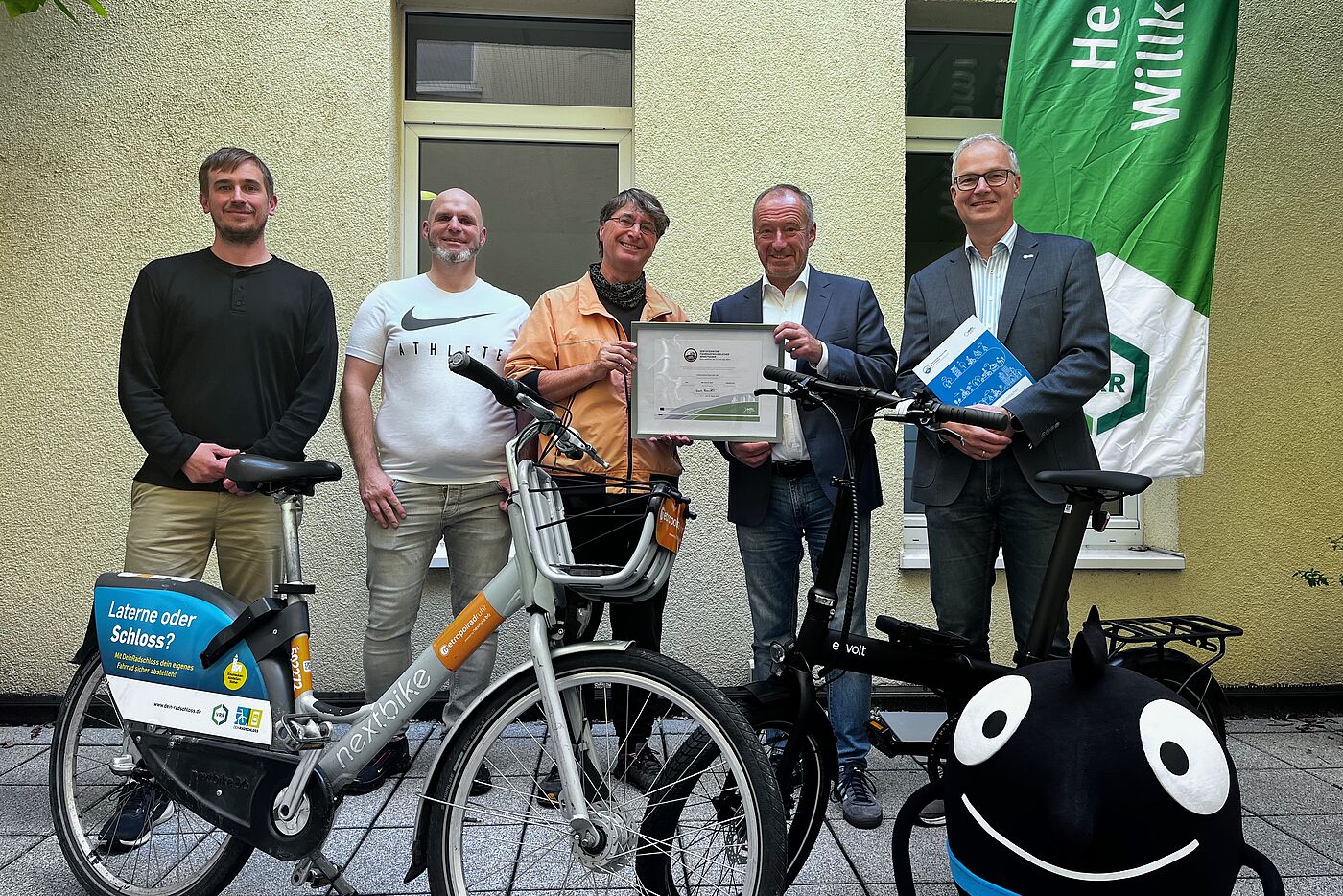 Zertifikatsübergabe Verkehrsverbund Rhein-Ruhr (VRR) 11.09.2025 v.l. Jan Gade und Stefan Oppermann (VRR Radverkehrskoordinatoren), Andreas K. Bittner (ADFC Projekt "Fahrradfreundlicher Arbeitgeber"), Oliver Wittke (VRR Vorstandssprecher) und Marc Zietan (ADFC NRW Landesvorstand) – im Vordergrund ein Metropolrad und rechts das Maskottchen der Europäischen Mobilitätswoche.